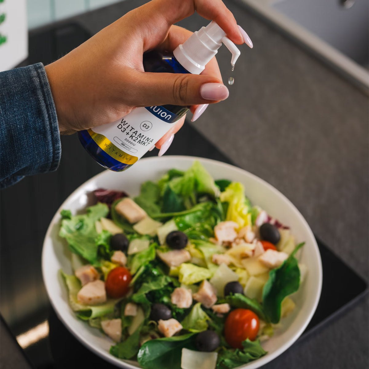 Eine hand hält eine Pipette des blauen Fläschchens von nuion über eine Schüssel mit frischem Salat, der mit Hähnchenstücken, grünen Blättern, schwarzen Oliven und Kirschtomaten belegt ist.