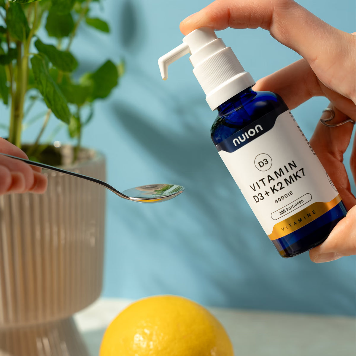 Eine Person hält einen Löffel in der Nähe eines blauen Fläschchens mit Flüssigkeit, das den Markennamen nuion und die Bezeichnung „Vitamin D3 + K2MK7 4000 IE“ von Puromedica trägt.