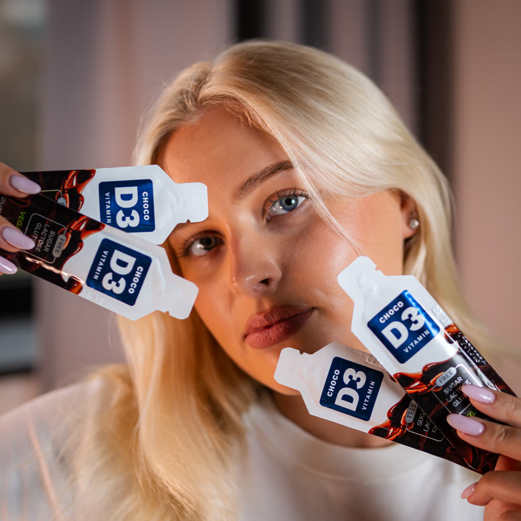 Eine junge Frau hält mehrere Packungen Vitamin D3 Schokolade von Puromedica in die Kamera. Sie hat lange, blonde Haare und trägt ein schlichtes, helles Oberteil, während sie mit einem neutralen Gesichtsausdruck in die Kamera blickt.