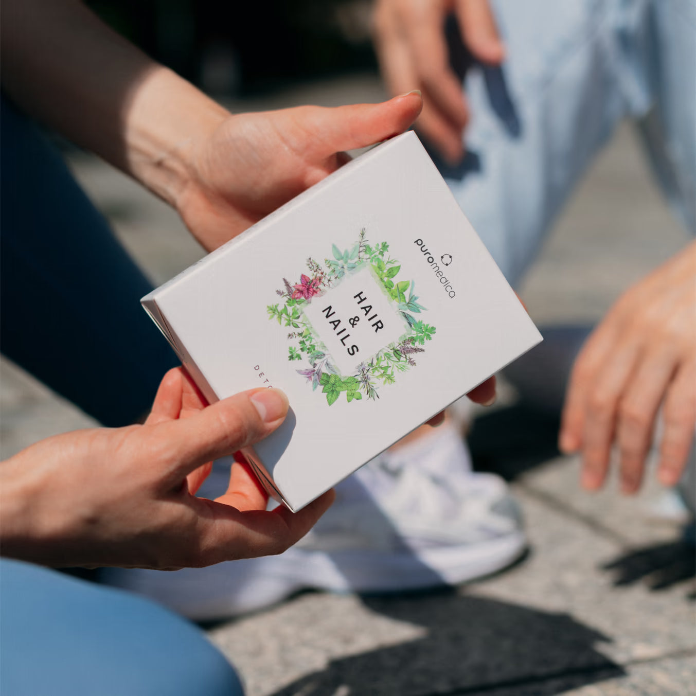 Eine Person hält eine weiße Box mit dem Aufdruck HAIR & NAILS in den Händen. Die Box gehört zu Puromedica und ist von einer Vielzahl von grünen Pflanzenmotiven umrahmt, während im Hintergrund eine weitere Person auf dem Boden sitzt.